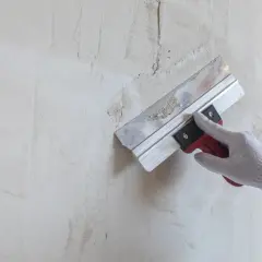 Ręka w rękawicy nakłada masę akrylową na pęknięcia na ścianie za pomocą szpachli.