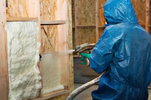 Osoba w niebieskim kombinezonie aplikuje izolację w sprayu na drewnianą konstrukcję ściany.