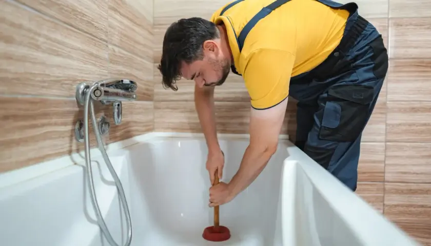 Mężczyzna w żółtej koszulce i granatowych ogrodniczkach używa przepychacza, by jak wyczyścić odpływ w wannie.