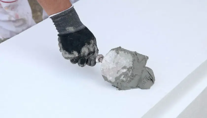 Ręka w rękawicy nakłada klej budowlany w piance na biały styropian.