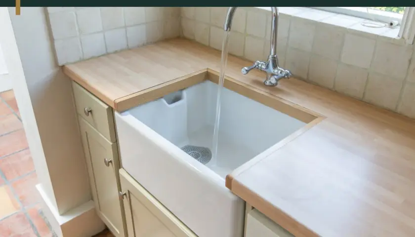 Biała, ceramiczna umywalka do kuchni z chromowaną baterią, w której płynie woda. Blat drewniany, szafki jasne.