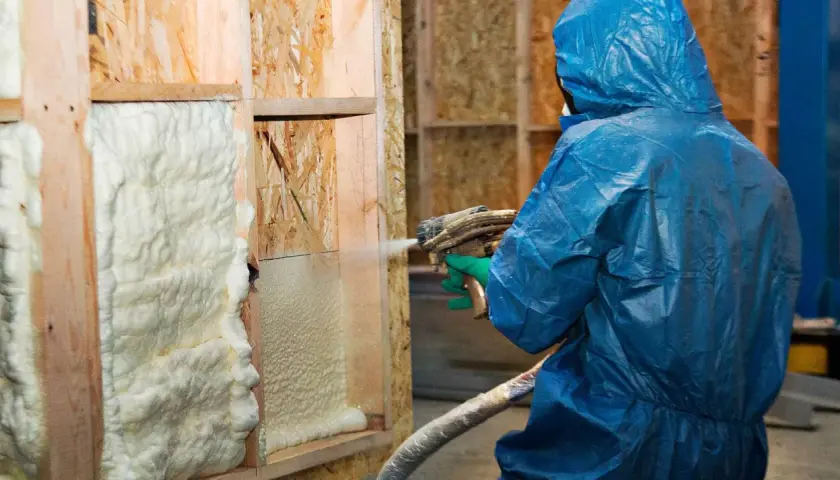 Osoba w niebieskim kombinezonie aplikuje izolację w sprayu na drewnianą konstrukcję ściany.