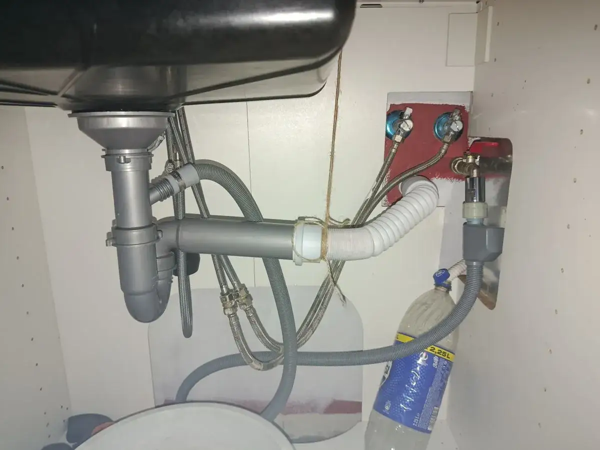 Pod zlewem widać instalację hydrauliczną, w tym syfon i rury. Wąż odpływowy jest podłączony do butelki, co sugeruje zapowietrzony odpływ zlewu.