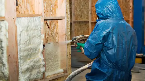 Osoba w niebieskim kombinezonie aplikuje izolację w sprayu na drewnianą konstrukcję ściany.